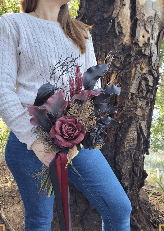 Artificial wedding bouquet close-up held at waist height, showing black and burgundy faux calla lilies and roses with bronze sprigs and long ribbons. Made in Melbourne by Flowers For Ever After, shipping Australia and Worldwide.