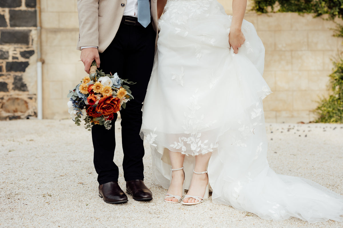 A close-up showing the lower half of a bride in a white lace-trimmed gown holding out her skirt, standing beside a groom in a beige blazer, black trousers, and brown shoes who is holding a colorful bouquet. Flowers by Flowers For Ever After