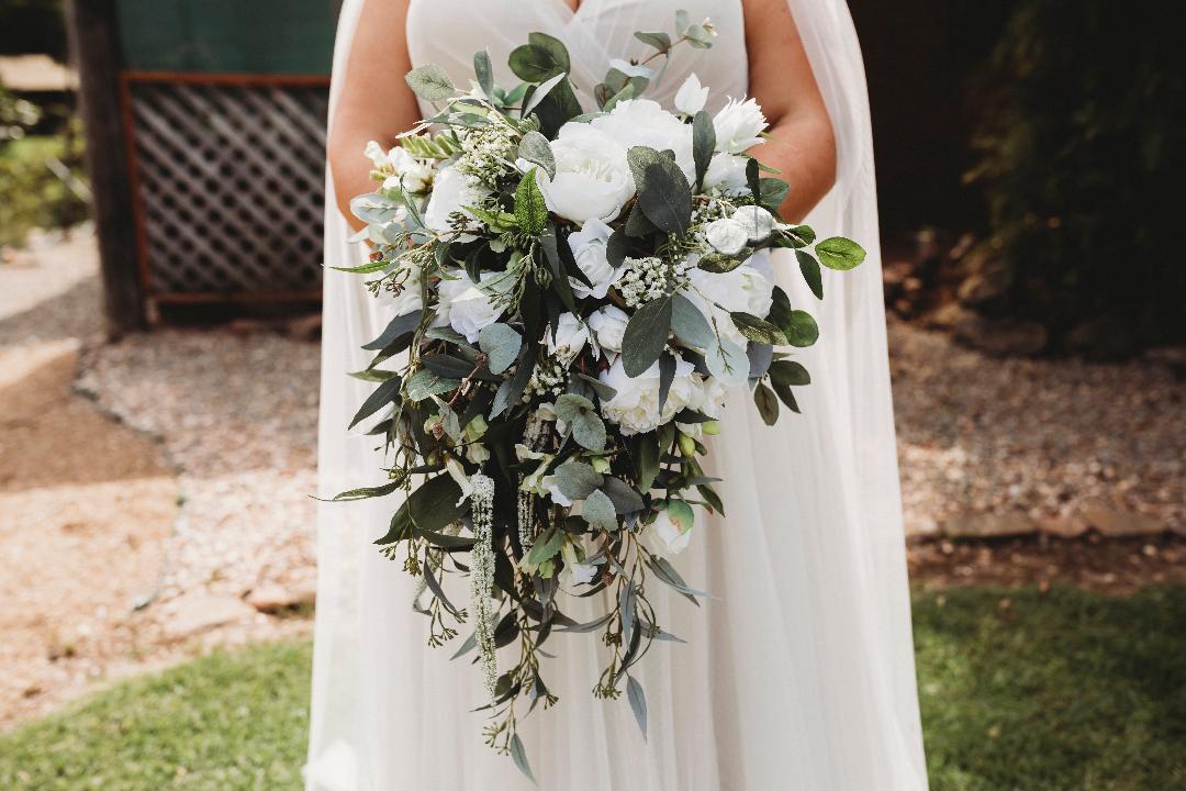 A bride in a white wedding gown holding a lush artificial bridal bouquet of white flowers and green foliage. Flowers by Flowers For Ever After