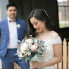 A bride in an off-white lace wedding dress and a man in a blue suit holding a bouquet, photographed outdoors with a blurred background.Flowers by Flowers For Ever After