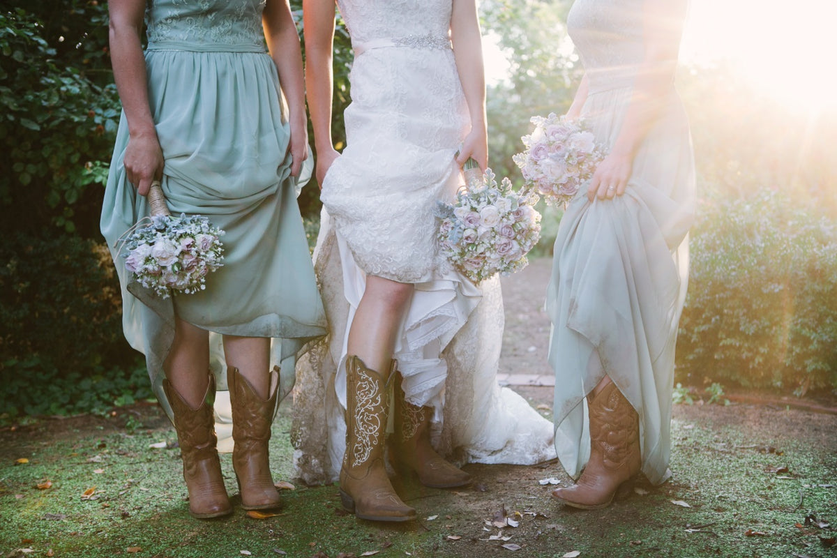Three women standing outdoors in wedding attire, shown from the waist down. Two wear sage green bridesmaid dresses and hold pastel flower bouquets, while the center woman wears a white lace wedding gown. Flowers by Flowers For Ever After