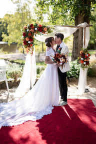 A bride in a long white lace wedding gown and a groom in a cream suit kiss under a rustic wooden arch decorated with white draped fabric and faux red, orange, and burgundy floral arrangements. Flowers by Flowers For Ever After
