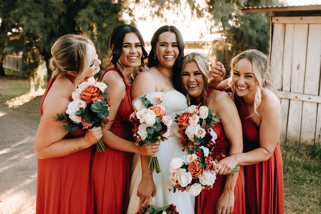 A bride in a white strapless gown surrounded by five bridesmaids in matching red dresses, holding orange, white, and red faux flower bouquets. Flowers by Flowers For Ever After