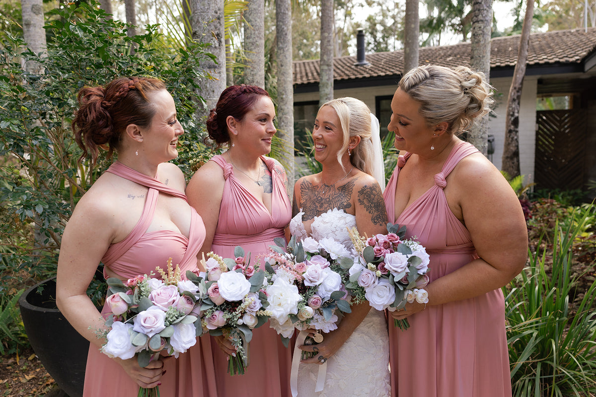 Four bridesmaids in matching dusty pink halter dresses stand outdoors with a bride in a white gown, holding pastel faux flower bouquets. Flowers by Flowers For Ever After