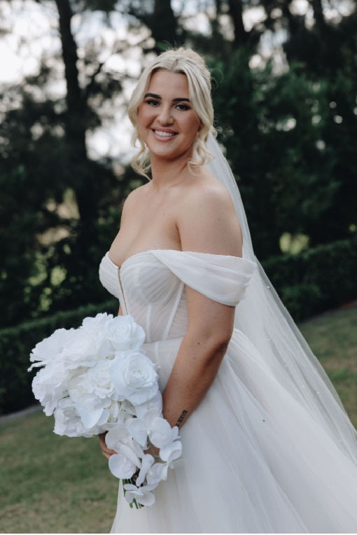 A bride in an off-the-shoulder white wedding gown with a long veil, holding a large white artificial floral bouquet outdoors in front of a blurred green garden background. Flowers by Flowers For Ever After