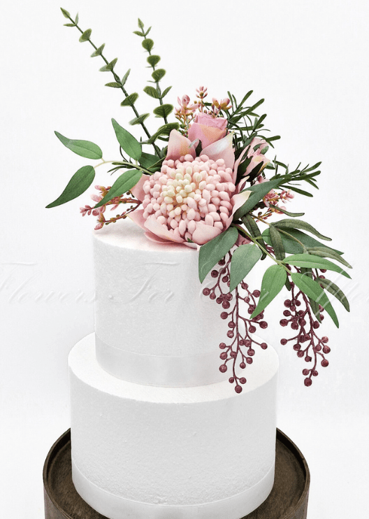 Artificial wedding cake decoration close-up with pink faux waratah blooms and green native-style foliage. For birthday, event and celebration cakes. Made in Melbourne by Flowers For Ever After, shipping Australia and Worldwide.