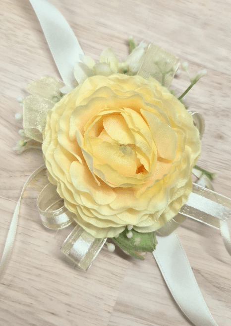 Artificial wedding wrist corsage, overhead view of a pale yellow faux ranunculus, pale green blossoms and gold-edged ivory ribbon loops. For formals, proms and ball occasions. Made in Melbourne by Flowers For Ever After, shipping Australia and Worldwide.