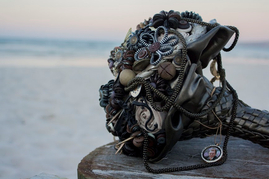 A handmade gothic-style button and beadbridal bouquet displayed on a wooden block with a blurred beach background. The piece features a dark brown and black buttons decorated with intricate silver and bronze filigree, beads, and draped chains with a small portrait pendant. Flowers by Flowers For Ever After