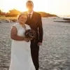 A bride in a white wedding gown and a groom in a dark suit standing together on a beach at sunset, holding a bouquet.