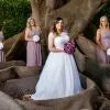 A bride in a white wedding gown holding a bouquet of purple flowers, standing outdoors next to bridesmaids in lavender dresses beside a large tree trunk.Flowers by Flowers For Ever After