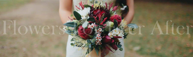 A close-up of a person holding a large, lush faux floral bouquet with deep red, burgundy, white, and green flowers, set against a softly blurred outdoor background. Flowers by Flowers For Ever After