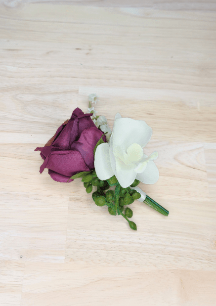 Artificial wedding boutonniere, overhead view of plum faux rosebud, white orchid and green berries. For formals, proms and ball occasions. Made in Melbourne by Flowers For Ever After, shipping Australia and Worldwide.
