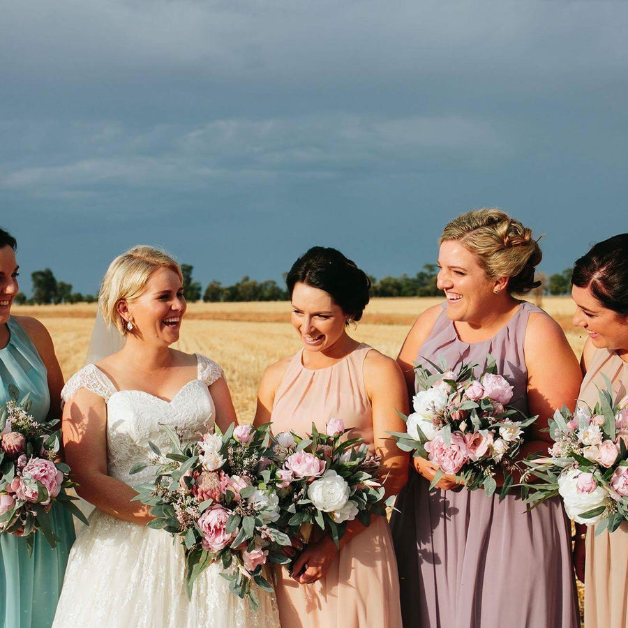 Five women standing together in a golden wheat field under a cloudy sky, wearing coordinated sleeveless dresses in aqua, white, blush, mauve, and beige, each holding a bouquets of artificial pink and white flowers, by Flowers For Ever After.