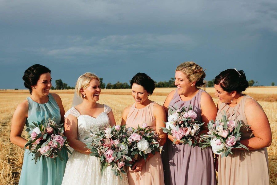 Five women standing together in a golden wheat field under a cloudy sky, wearing coordinated sleeveless dresses in aqua, white, blush, mauve, and beige, each holding a bouquets of artificial pink and white flowers, by Flowers For Ever After.
