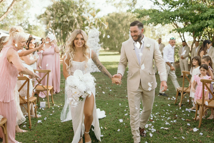 A bride in a white sleeveless gown holding a large faux white floral bouquet walks hand in hand with a groom in a light beige suit through a grassy outdoor ceremony aisle. Flowers by Flowers For Ever After